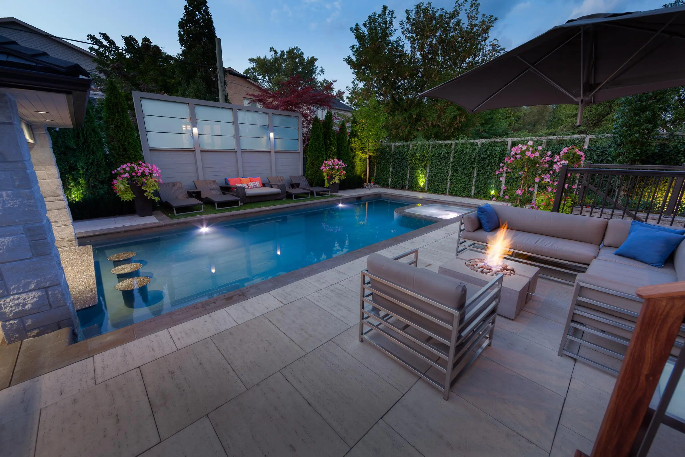 A lit square, modern concrete fire pit from Dekko’s Bravo collection forms the centerpiece of this modern patio corner, located to the right of the pool.